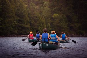 Group of kayakers paddle across a dark lake in colorful life jackets with forest backdrop at dusk.
