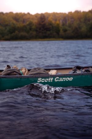 A green Scott canoe floats on rippling lake water with forest shoreline visible in the background at dusk.