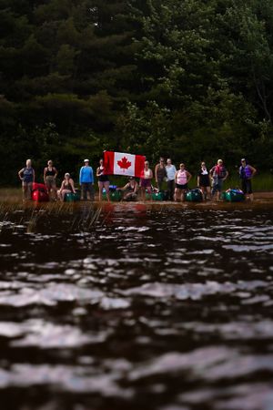 A group of people gathered at a lakeside shore with a Canadian flag during a summer gathering.