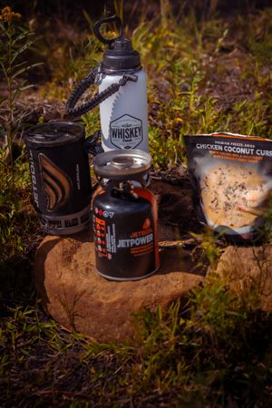 Camping supplies including a water bottle, JetPower fuel canister, and snacks arranged on a rock in nature.