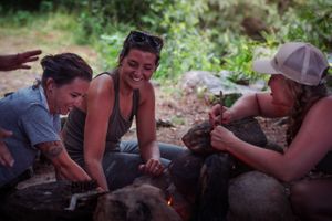 Three people gather around a campfire outdoors, sitting on logs and sharing a fun moment in nature together.