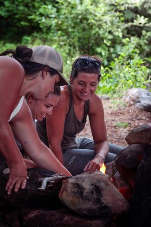 Friends gather around a campfire outdoors, sharing laughter and warmth in a natural setting surrounded by greenery.