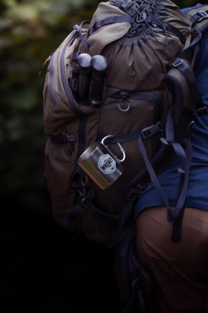 Close-up view of brown hiking backpack with gear and patches attached against dark background.