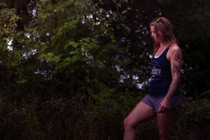 A figure in a dark tank top and shorts poses against greenery in moody evening lighting.