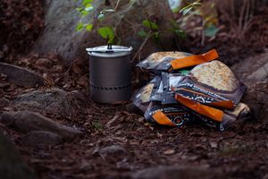Backpacking gear including a black camping stove and orange gloves resting on dark soil in an outdoor setting.
