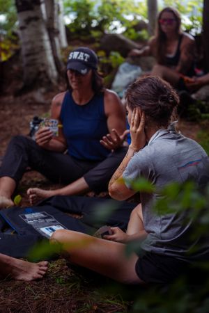 A group plays card games while sitting on the ground outdoors surrounded by trees and foliage.