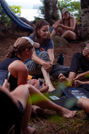 A group of people relax outdoors on forest ground while playing cards and having drinks together.