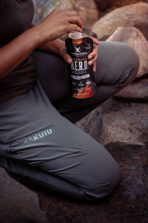 Person in gray Kuiu pants holds a Zero Energy drink while sitting on rocks outdoors.