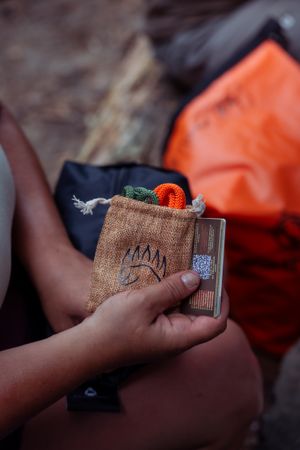 Hands hold a metal flask with brown burlap cover featuring a bear paw design next to an orange backpack outdoors.