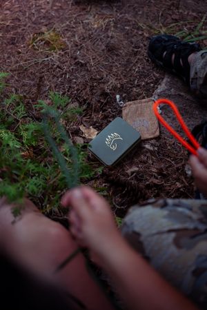 A dark leather notebook and red carabiner lie on forest ground covered in mulch and greenery.