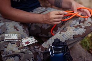 Hands holding bright orange paracord with camping gear and camouflage pants visible in background.
