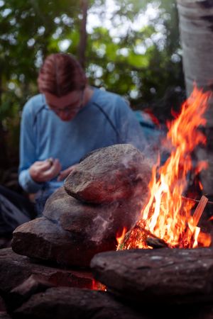 A bright campfire burns against stacked rocks while someone sits in the background during twilight.