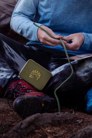 A pair of hands connect a green charging pack to devices while camping, with red hiking boots visible in the frame.