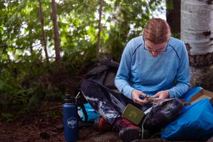 Someone in a blue sweater sits on the ground reviewing hiking gear with a water bottle and backpack nearby.