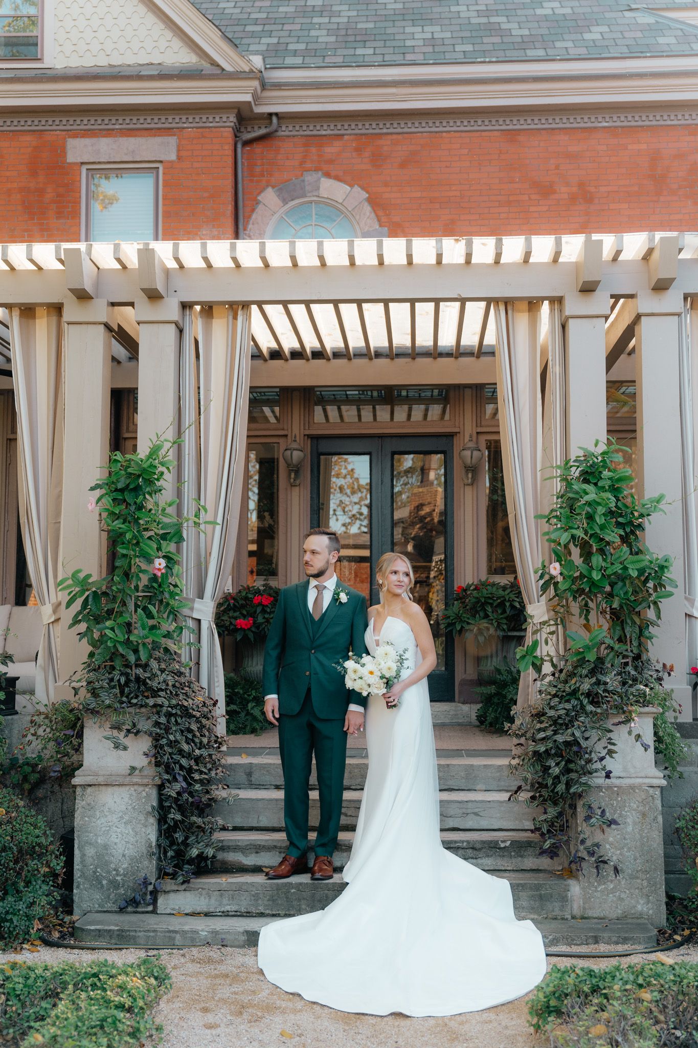 A wedding couple stands under an elegant pergola with white curtains and climbing vines at a historic brick mansion.