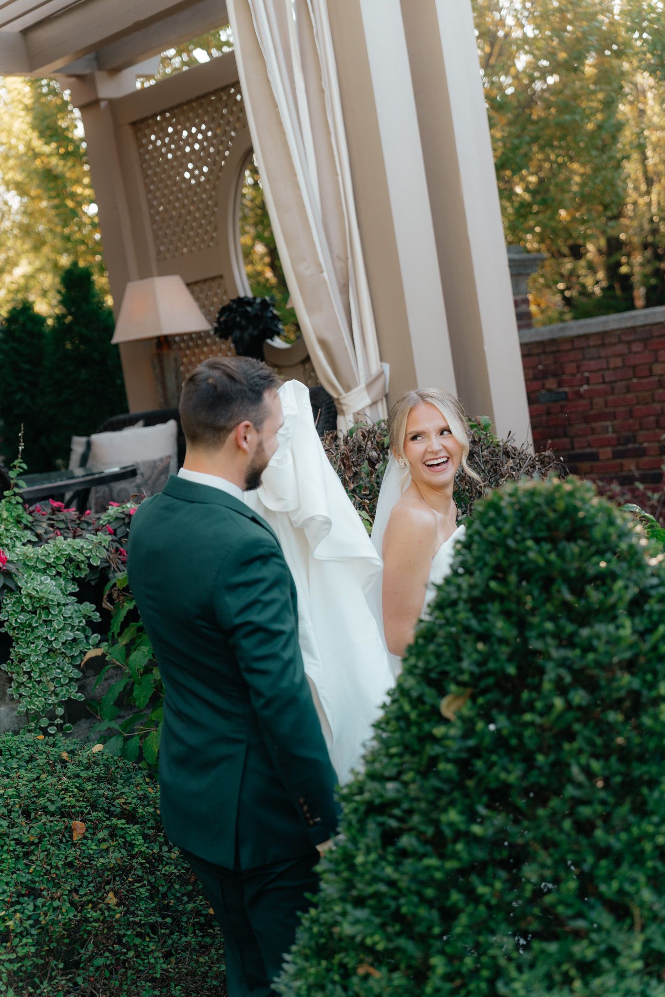 A couple in wedding attire share a joyful moment outdoors among manicured bushes and garden decor.