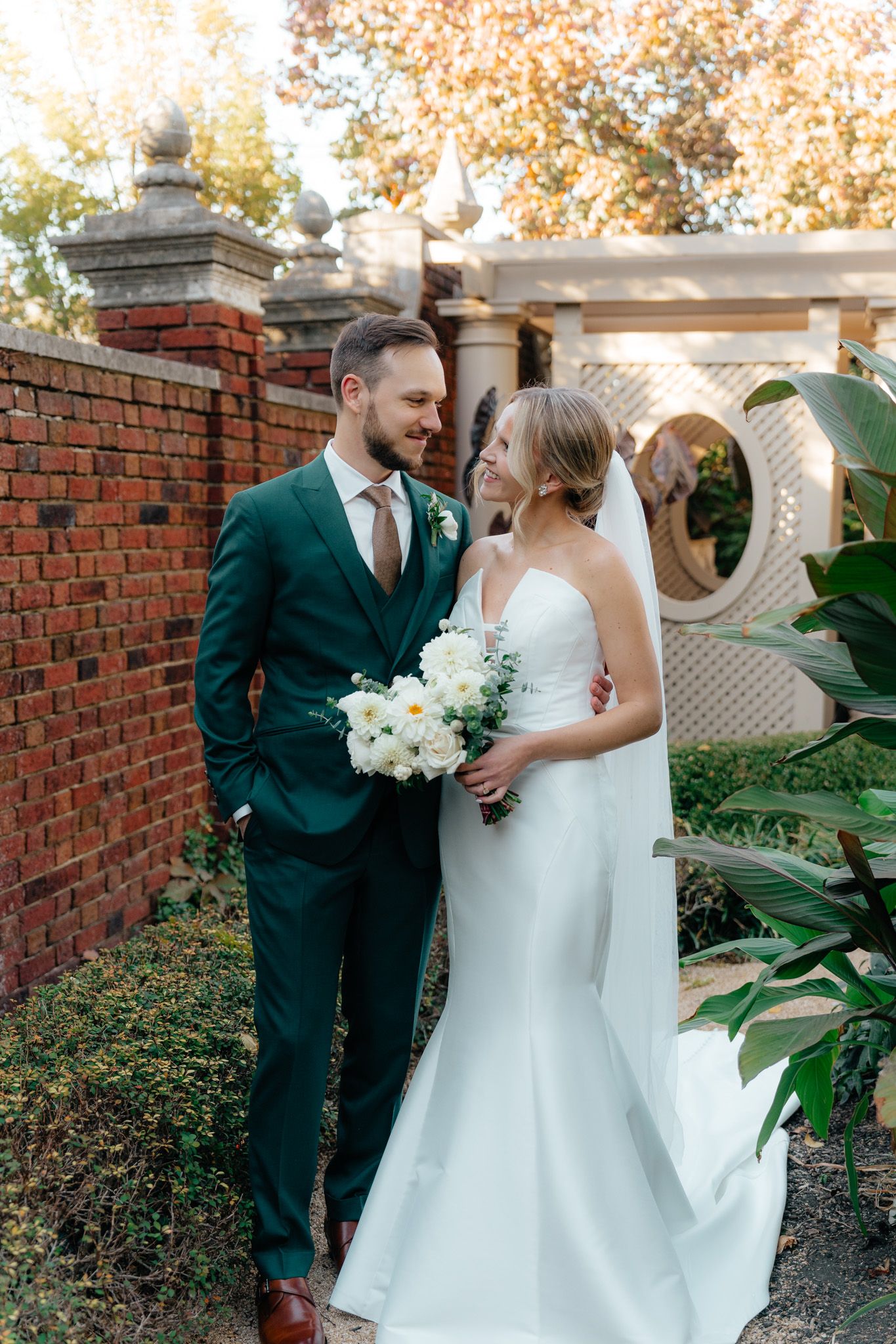 A couple in emerald green suit and white wedding gown pose in a garden setting with white flowers and brick walls.