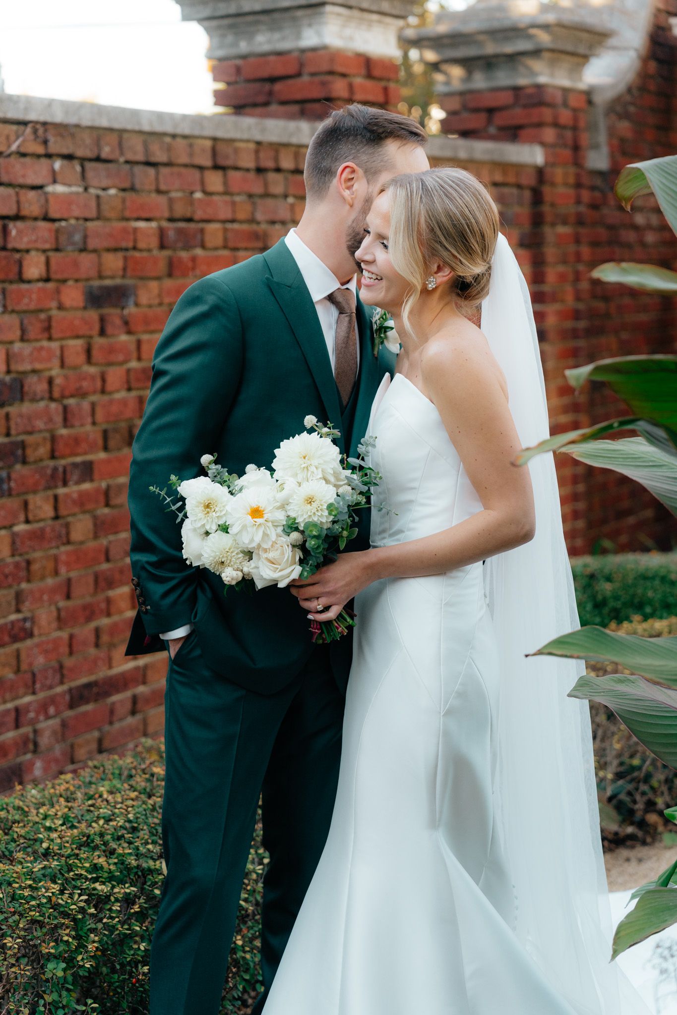 A romantic wedding moment against a brick wall backdrop with a classic white bridal bouquet and emerald green suit.