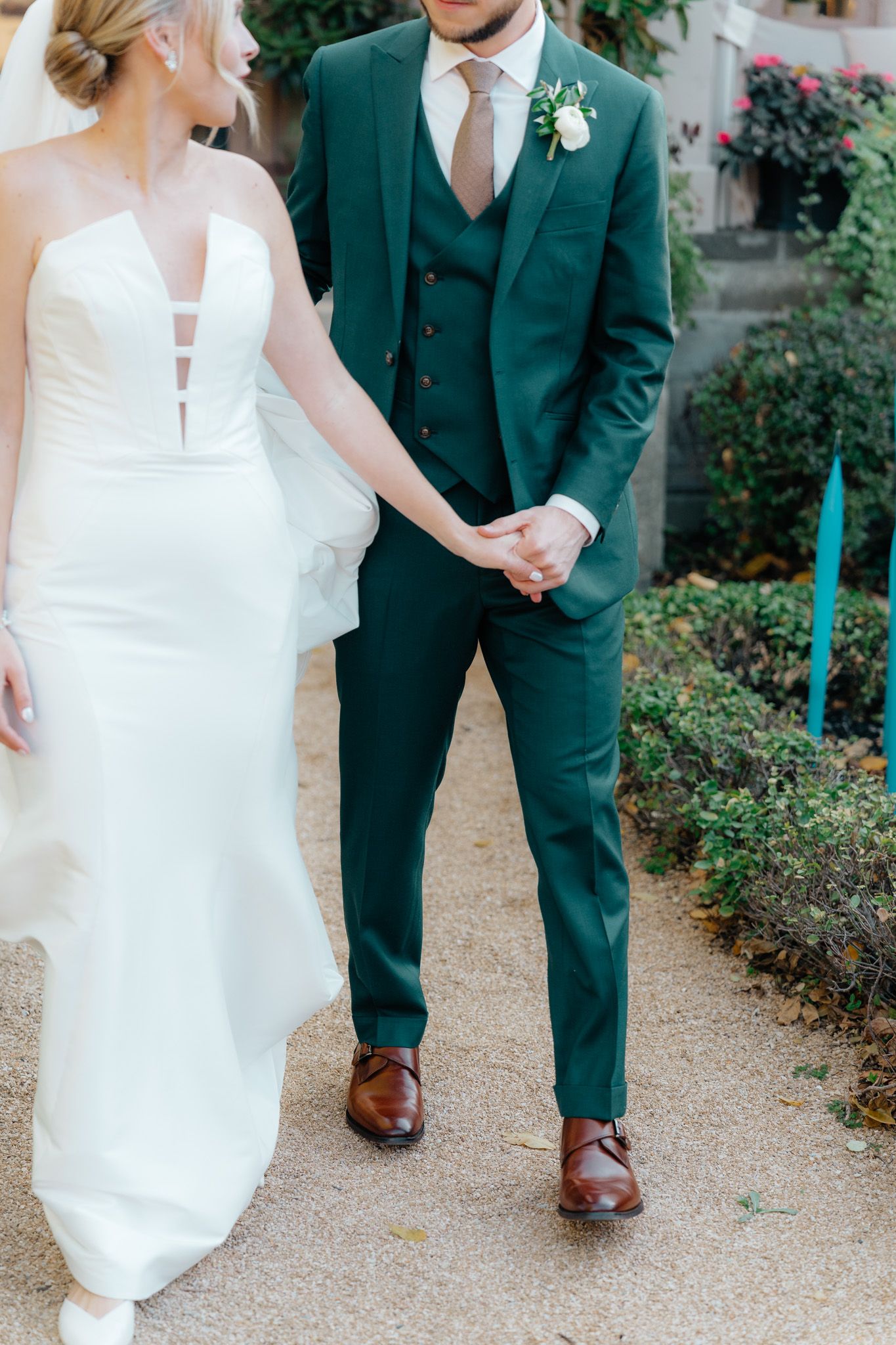 A couple in wedding attire holds hands, showcasing an emerald green three-piece suit with brown leather shoes and white gown.
