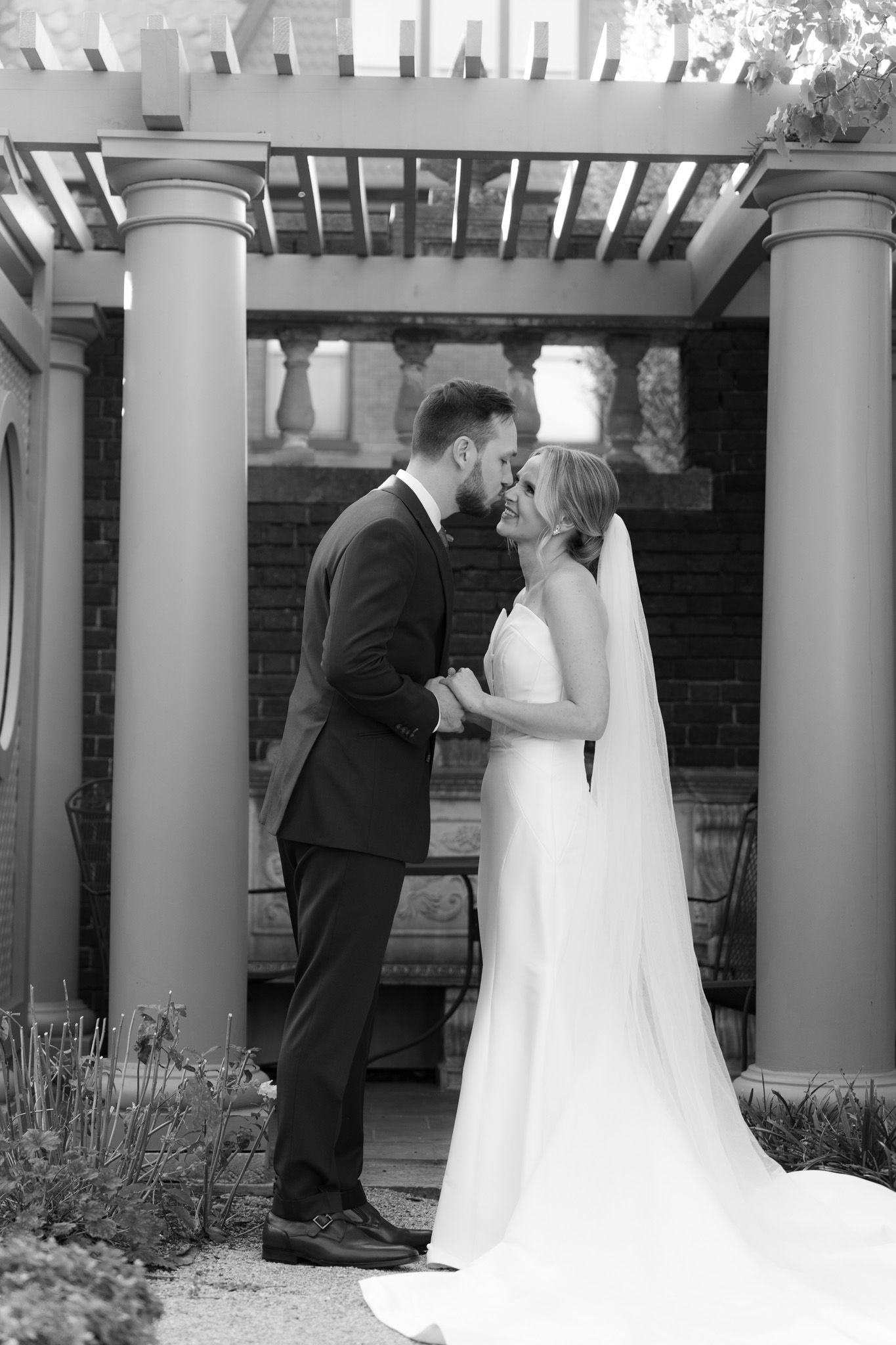 A romantic moment captured in black and white under a garden pergola with white pillars during a wedding ceremony.
