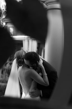 Romantic black and white wedding photo captured through a window frame showing a tender moment between newlyweds.
