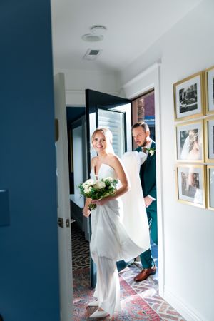 A couple in wedding attire enters through a doorway into a room with framed photos on white walls.