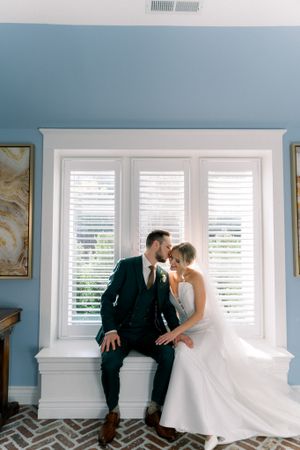 Wedding couple sits together on indoor window ledge with white plantation shutters and blue walls.