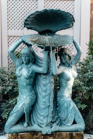 A blue-patina garden fountain sculpture featuring figures holding up a large leaf or shell-like structure as water spouts.