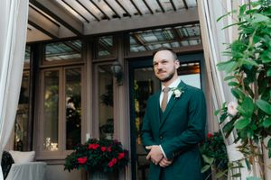 A well-dressed person in an emerald green suit stands near glass doors under a covered entryway with plants.
