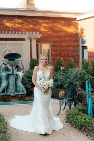 Wedding portrait in front of brick wall courtyard with garden statues and lush greenery.