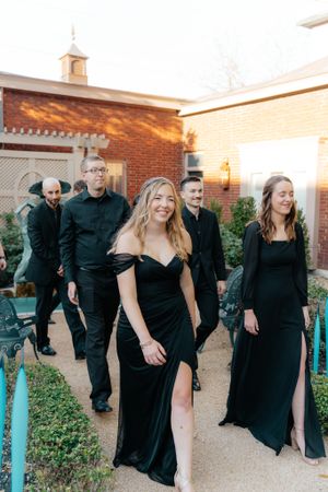 Wedding party members in elegant black attire walking along a brick building pathway at sunset.
