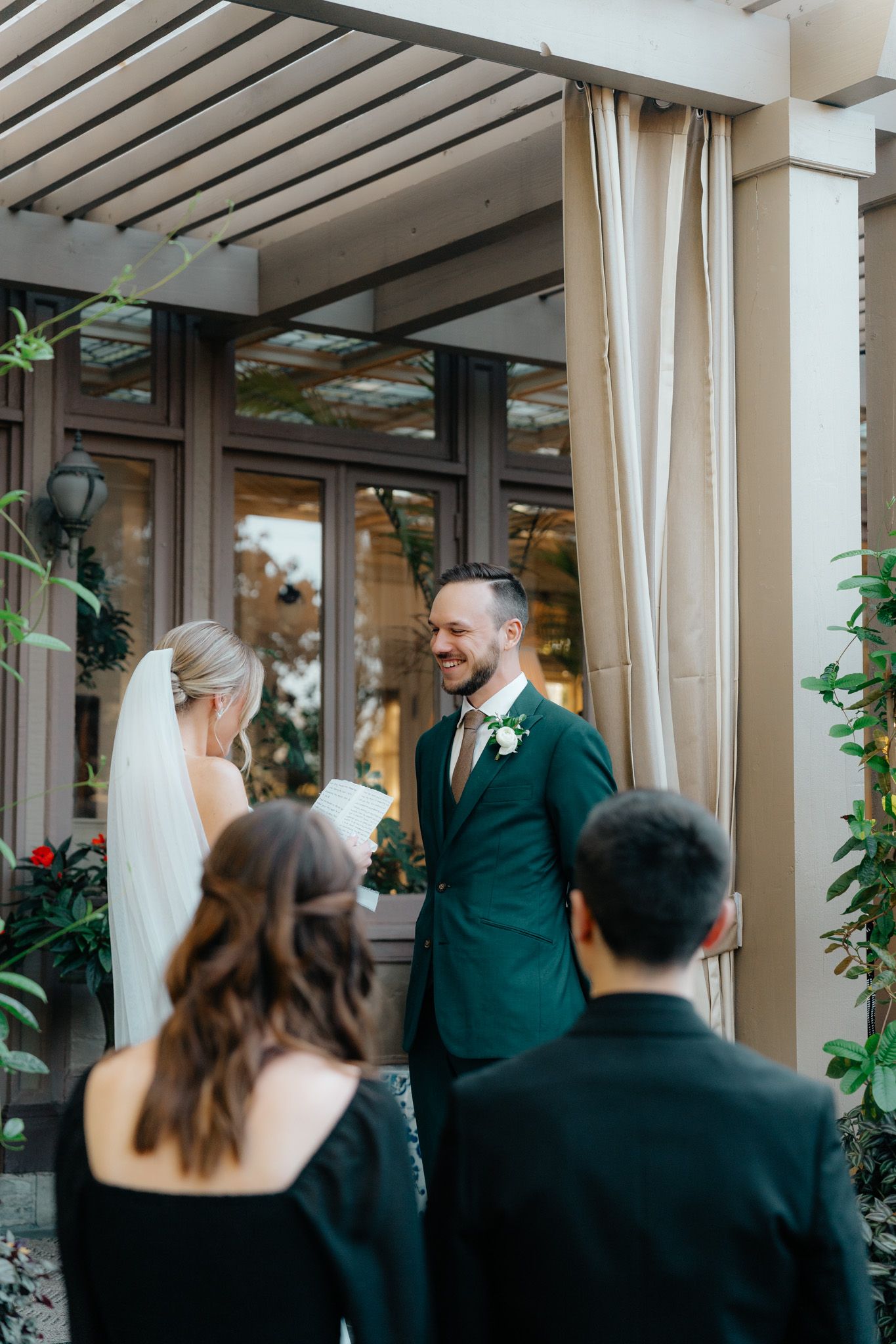 A wedding ceremony takes place under a covered patio area decorated with plants and curtains.