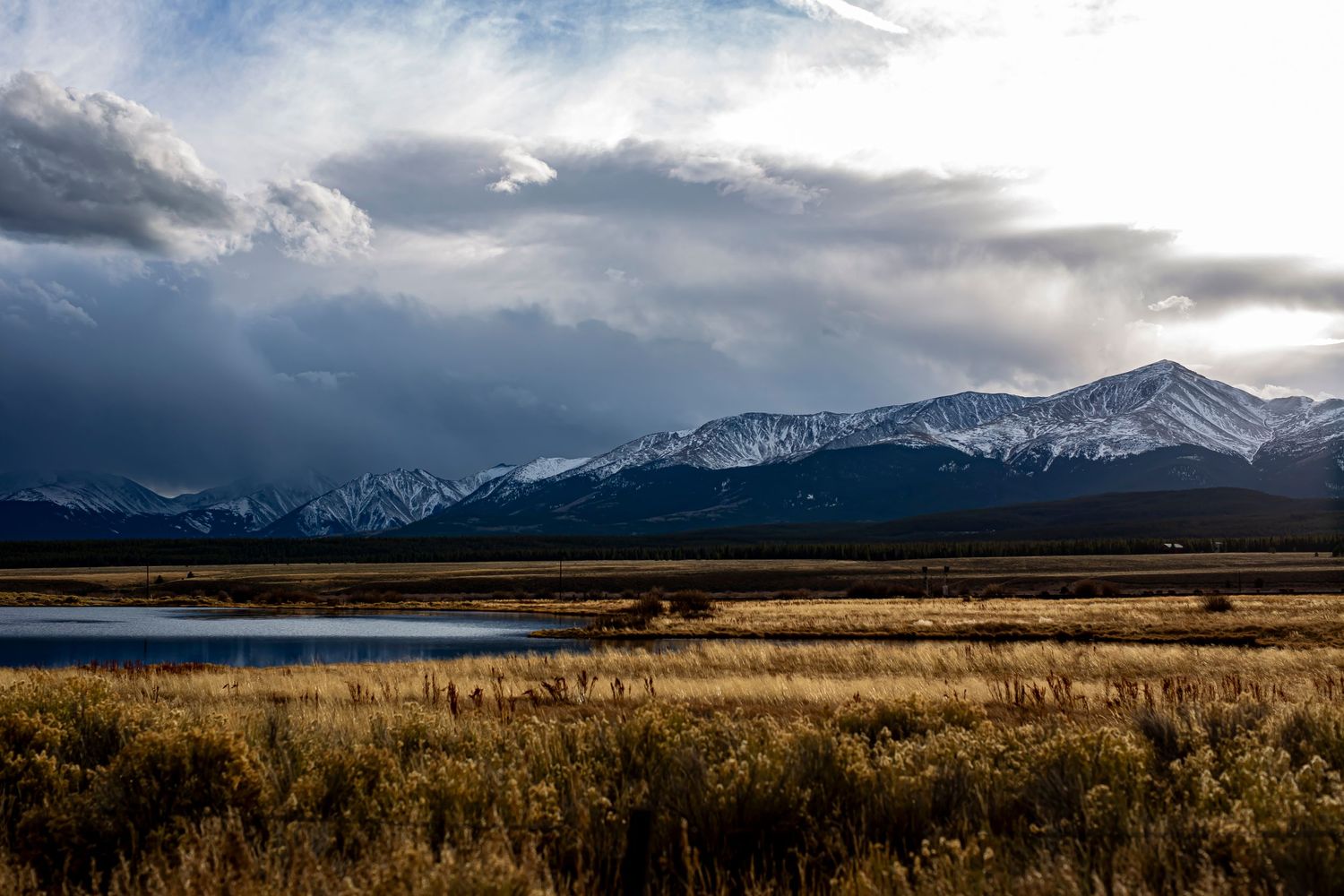 Snow-capped mountains rise behind a serene wetland landscape under dramatic cloudy skies.
