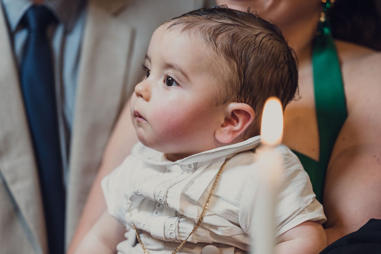 A baby in a white frilly dress is held at an outdoor celebration while guests gather around.