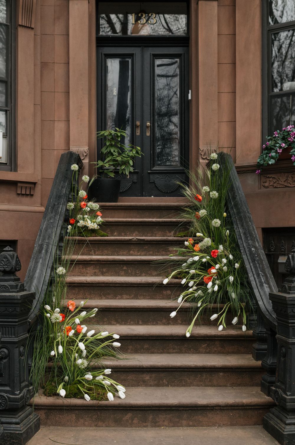 Stone steps leading to a black front door of a brownstone building decorated with red and white tulips along the railings.