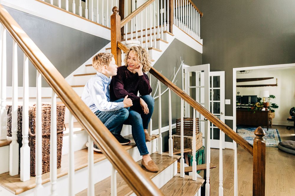A person sits with her son on wooden stairs in a home while scrolling on their phone in a casual indoor setting.
