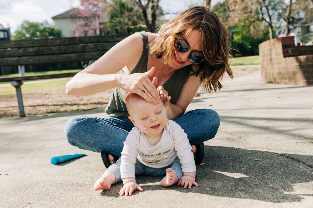 A playful moment outdoors on a sunny day as a baby explores the ground while wearing a white onesie and mom puts sunscreen on them