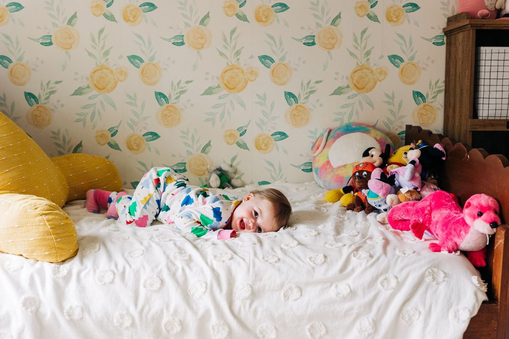 Child smiling and laying on white bed with colorful stuffed animals and floral wallpaper in background.