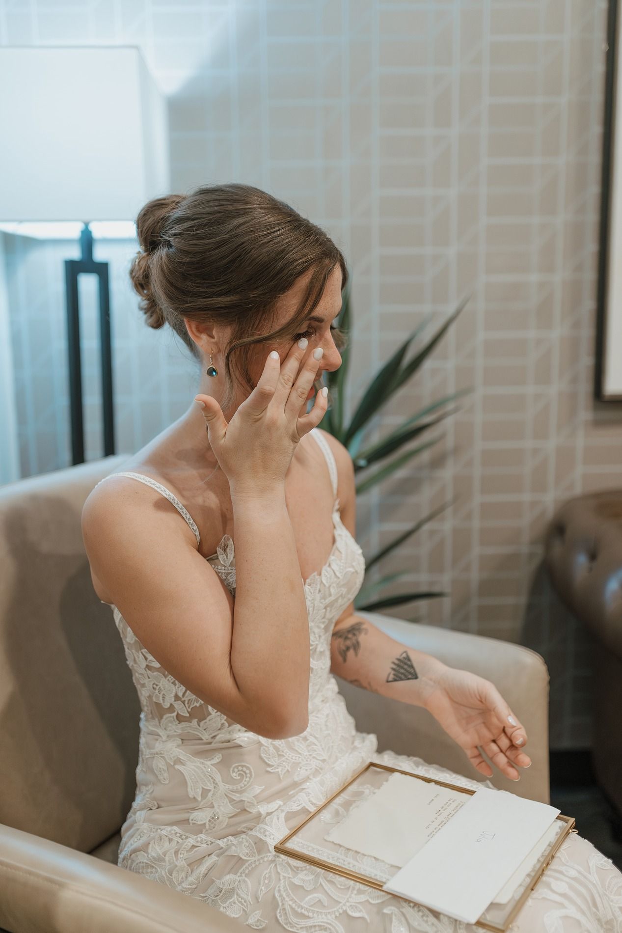 Emotional bride in white lace dress wipes tears while reading letter in modern interior setting.