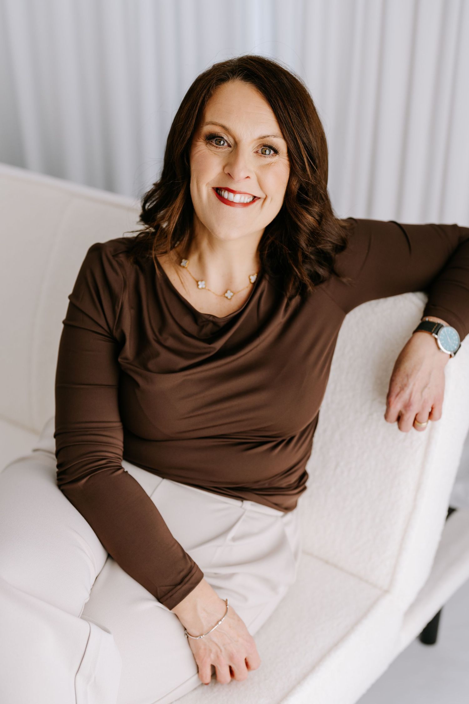 Professional portrait of someone in a brown cowl neck top sitting comfortably on a white couch against a light background.