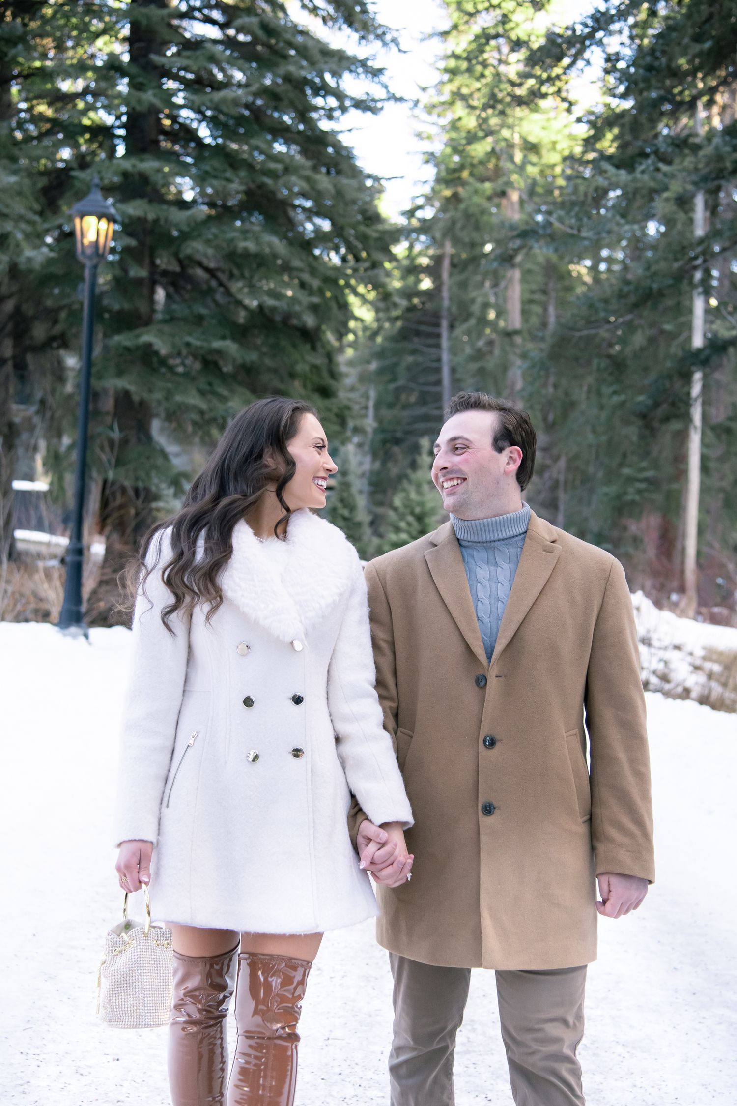 Couple in chic winter attire walking together through snowy forest path wearing white and tan coats for winter engagement in Vail Colorado
