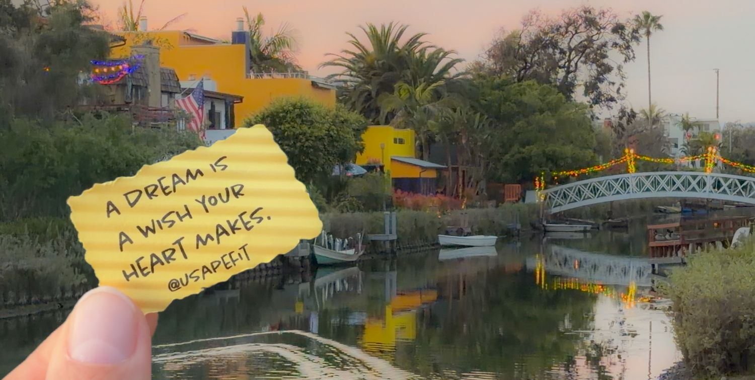 Hand holds yellow sticky note against backdrop of Venice canals and colorful buildings at sunset.