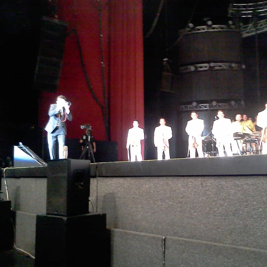 Performers on stage during a live show with red curtain backdrop and dark lighting.