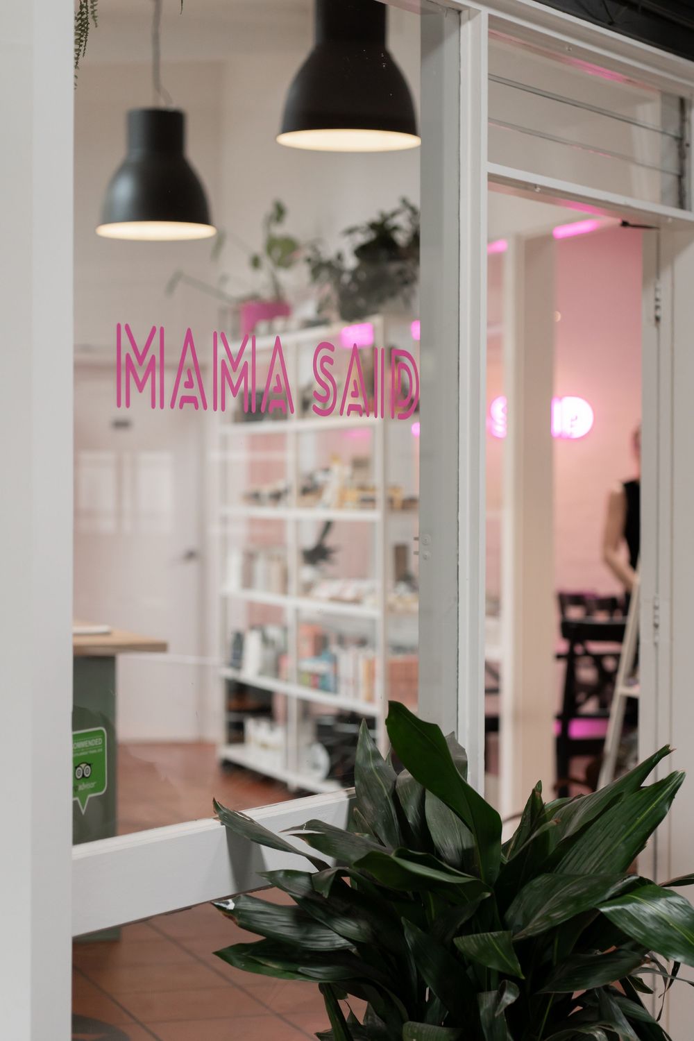 Modern cafe storefront with pink neon 'MAMA SAD' sign, black pendant lamps, and lush green plants in the entrance.