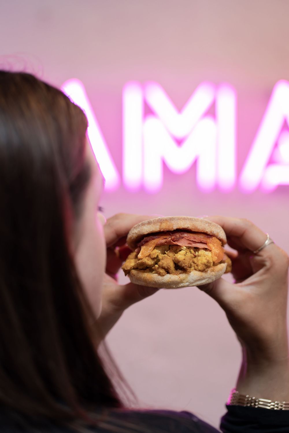 A person takes a bite of a chocolate chip cookie with a pink neon sign glowing in the background.