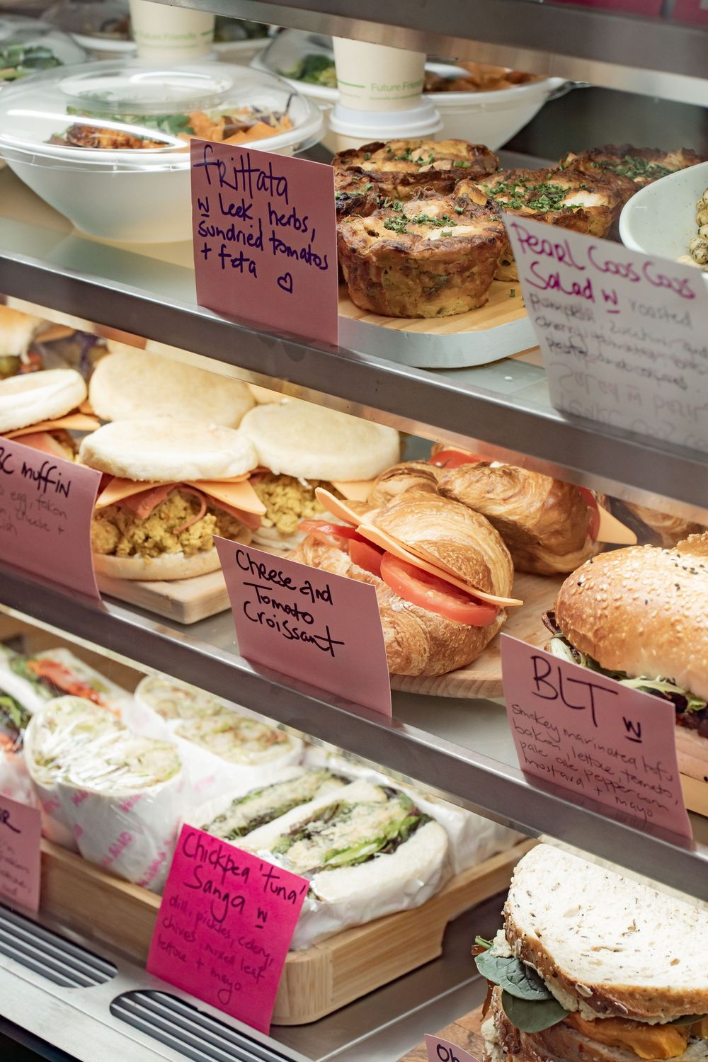 Display case filled with fresh sandwiches and baked goods with handwritten price tags on pink labels.