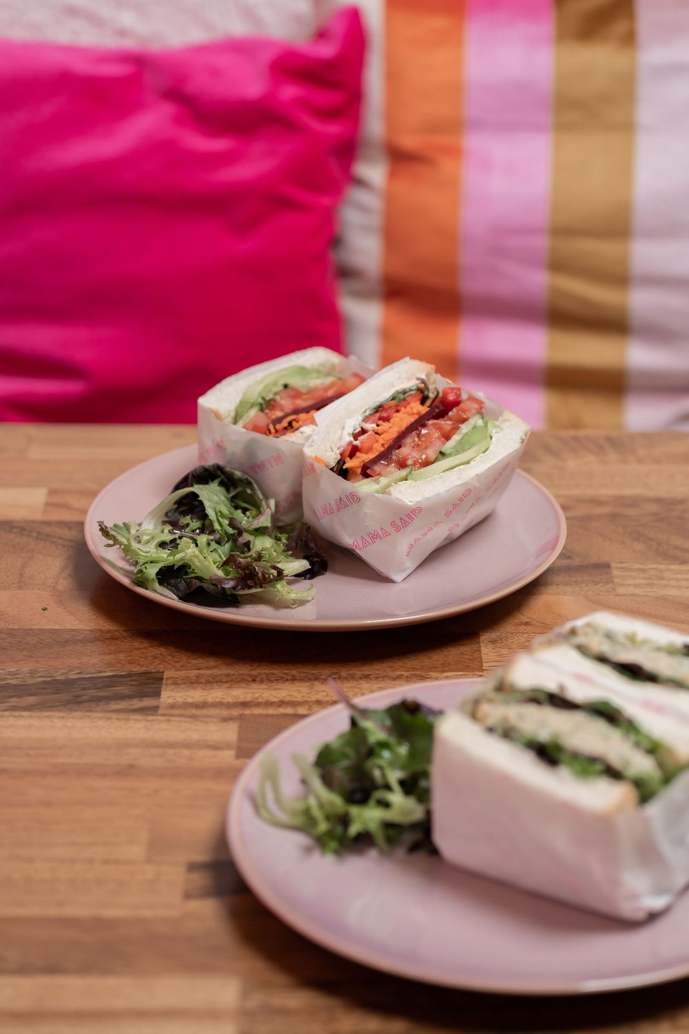Two wrapped sandwiches with fresh greens served on pink plates against a colorful striped background.