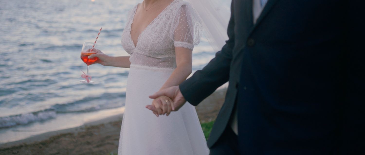 A couple in wedding attire holds hands by the water at sunset, with the bride wearing a white lace dress.