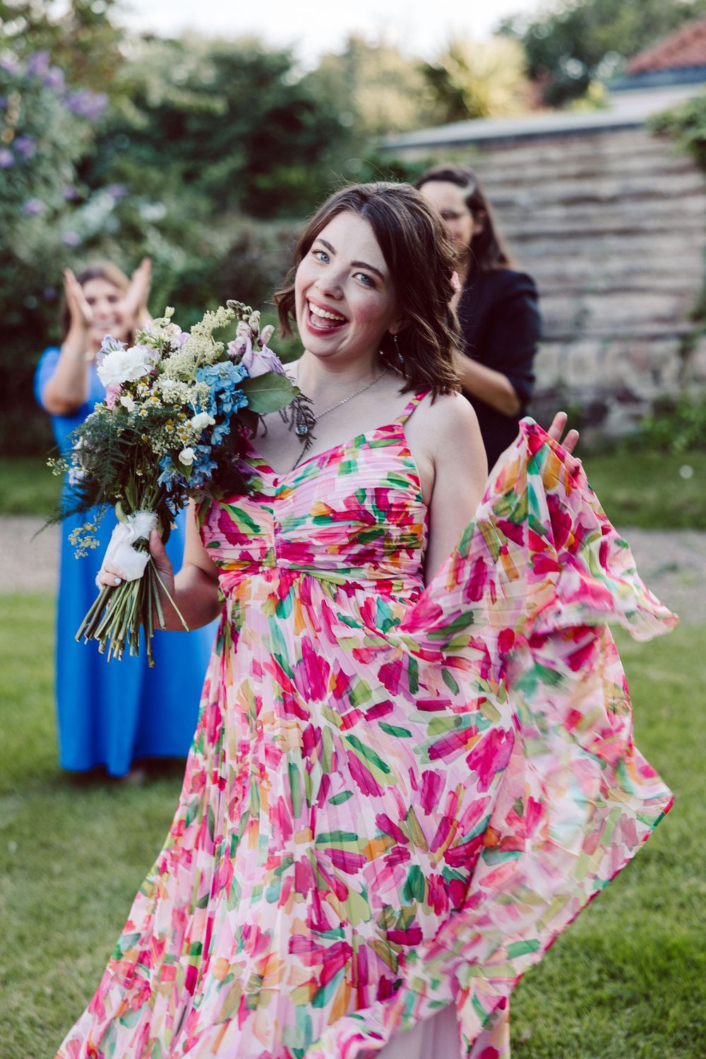 A floral print bridesmaid dress in pink and white swirls elegantly against a garden backdrop with blue-dressed attendants.