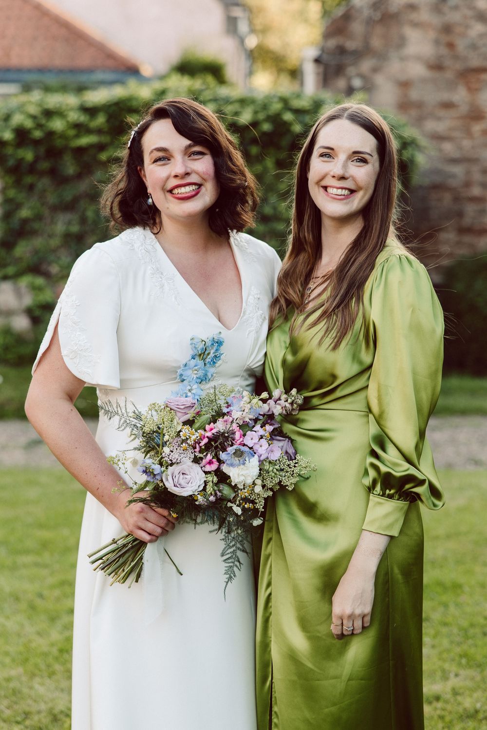 Two people in contrasting white and lime green dresses stand together holding a purple and white floral bouquet.